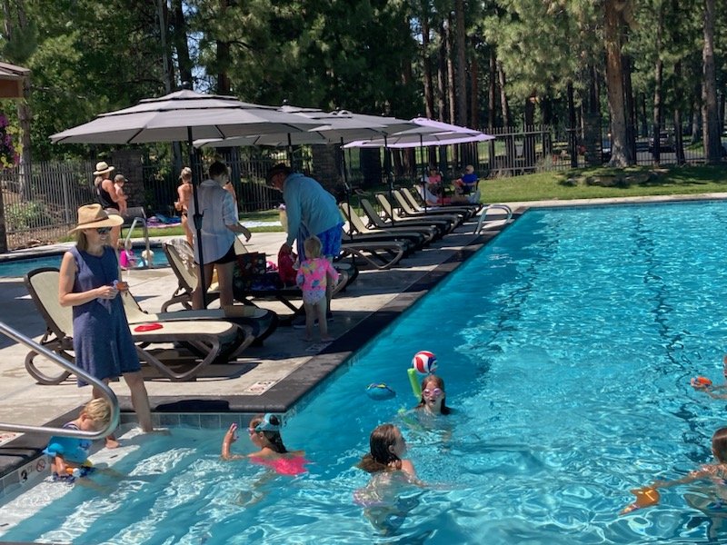 People enjoying a sunny day at an outdoor swimming pool, with children swimming and playing with a ball, and adults sitting on lounge chairs under umbrellas.