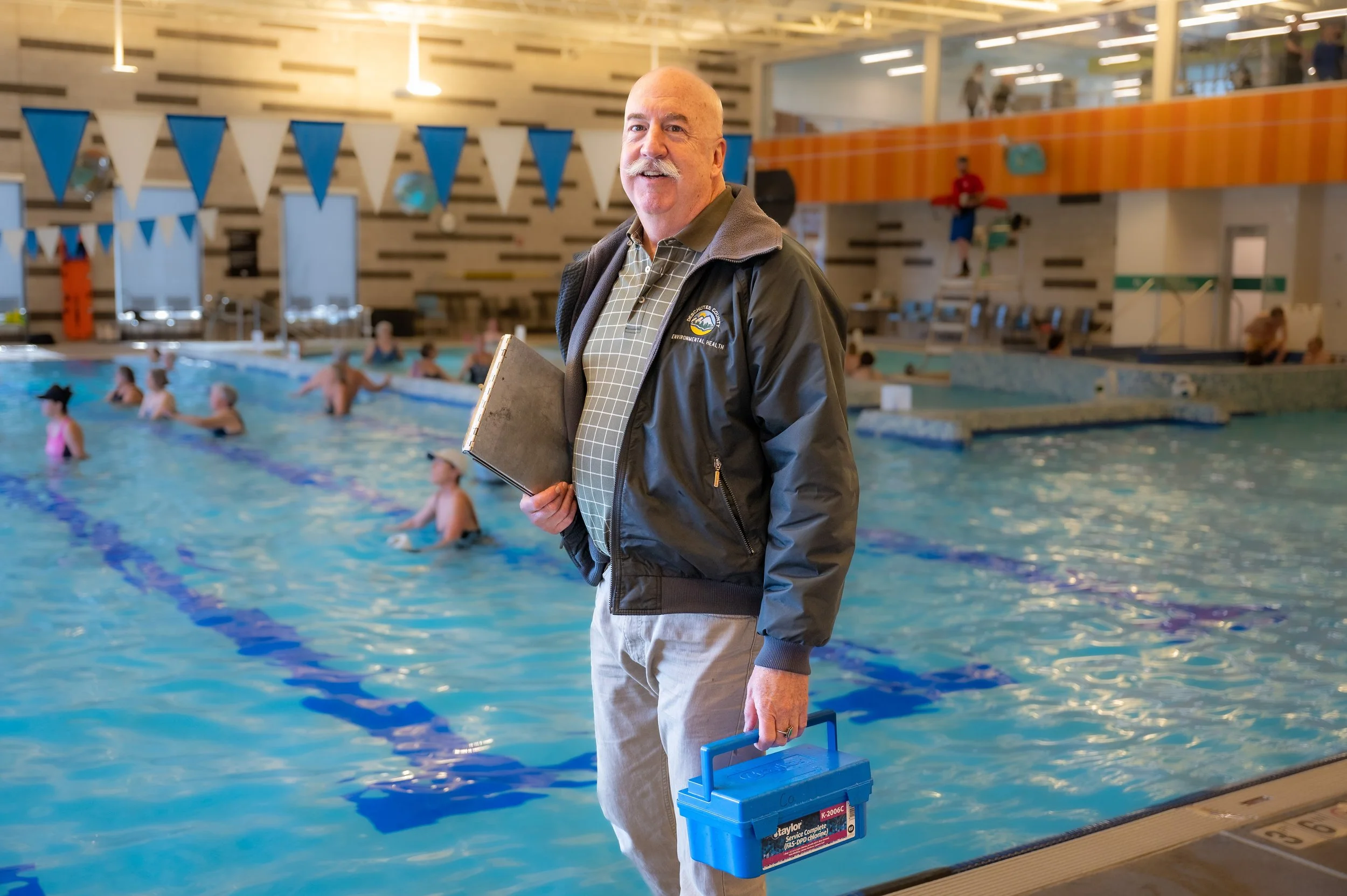 Lifeguard standing at the edge of an indoor swimming pool, holding a first aid kit and a rescue tube, with swimmers in the water behind him.