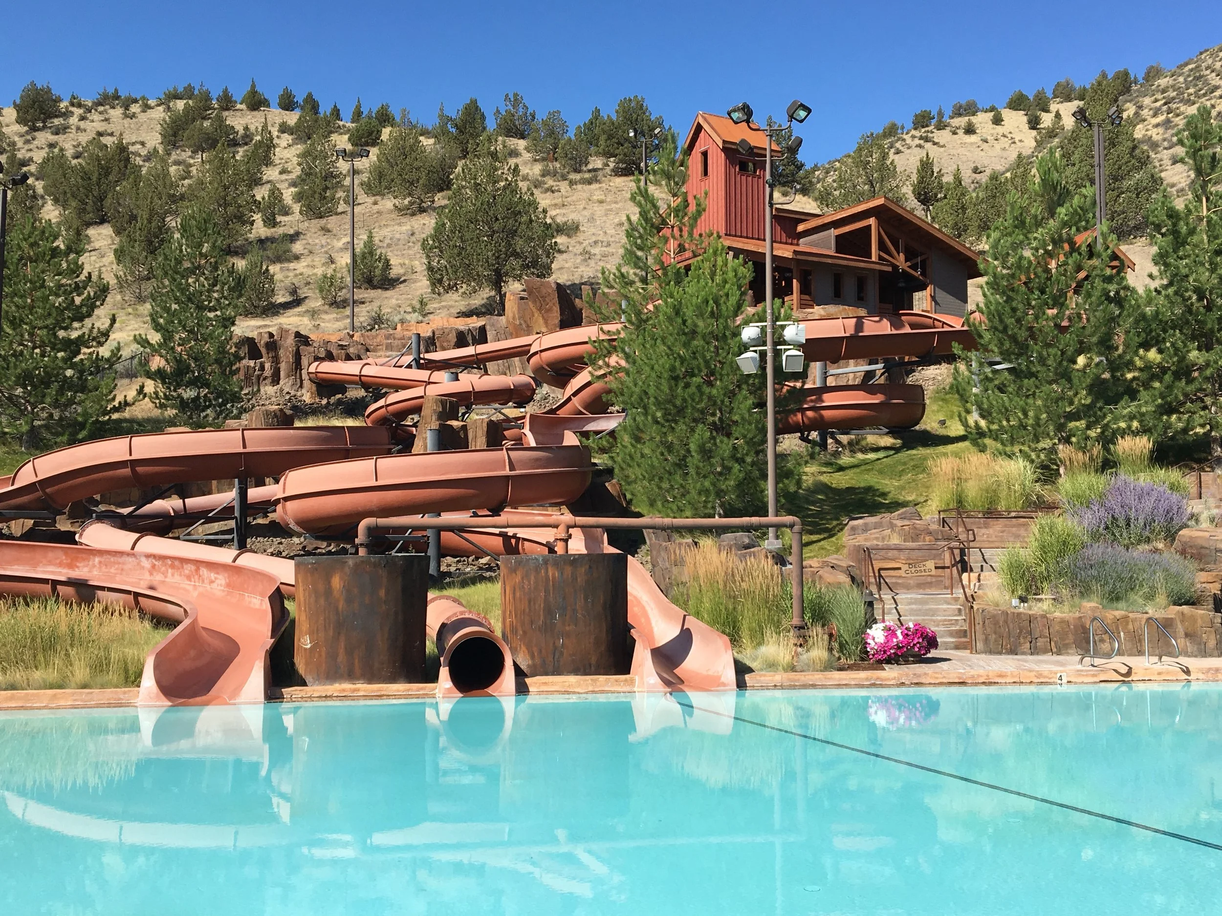 An outdoor water park pool with a large water slide in the background, surrounded by trees and mountainous terrain. The slide is pinkish-brown and has multiple twists and turns, with a small building at the top of the slide.