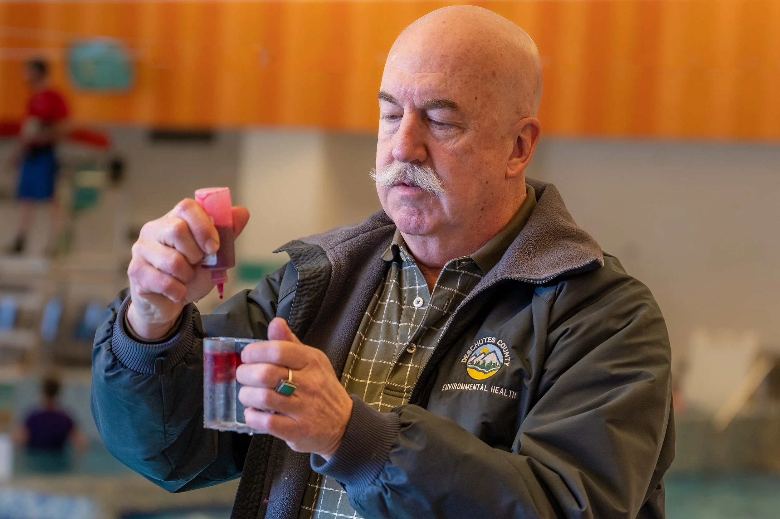 A man with a bald head and mustache wearing a jacket with a Deschutes County Environmental Health logo, filling a small clear container with red liquid from a dropper at an indoor facility.