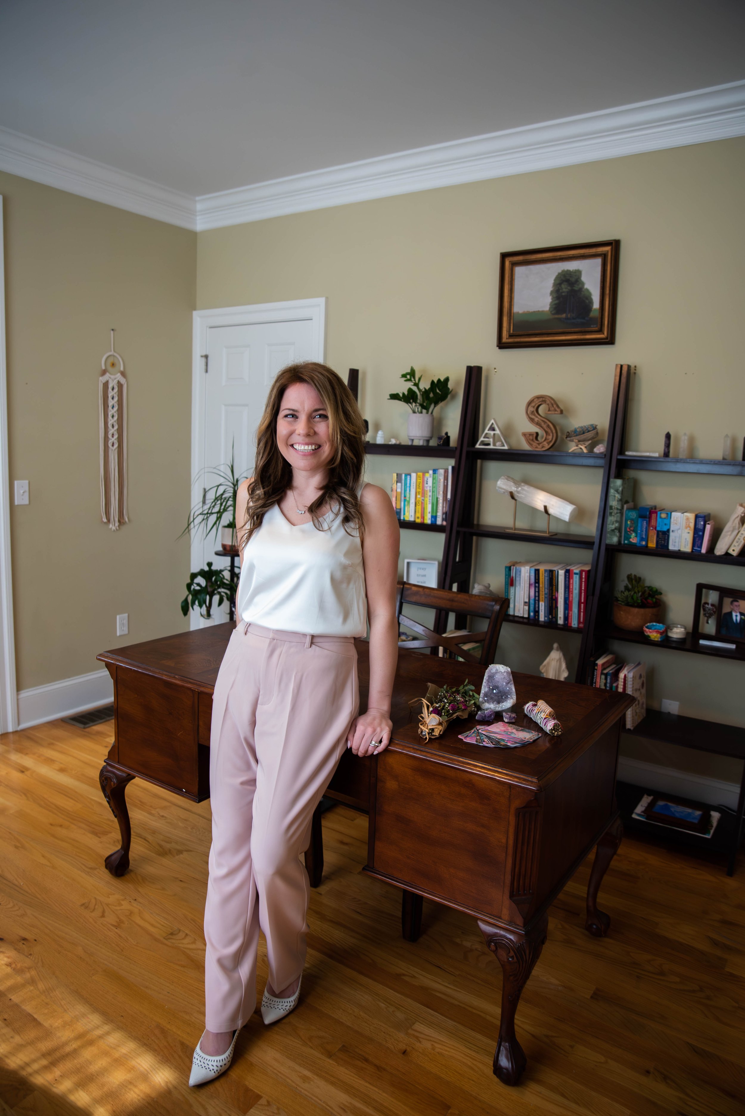 A woman smiling and standing near a wooden desk in a living room with bookshelves, plants, and decorations.