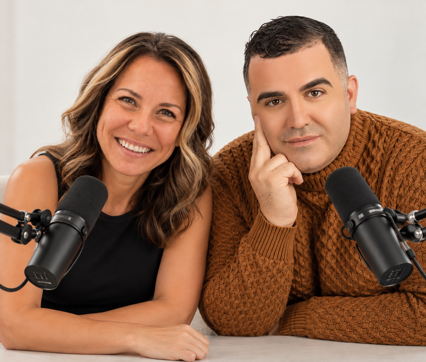 A smiling woman and a serious man sit at a table with microphones, appearing to be recording a podcast or radio show.