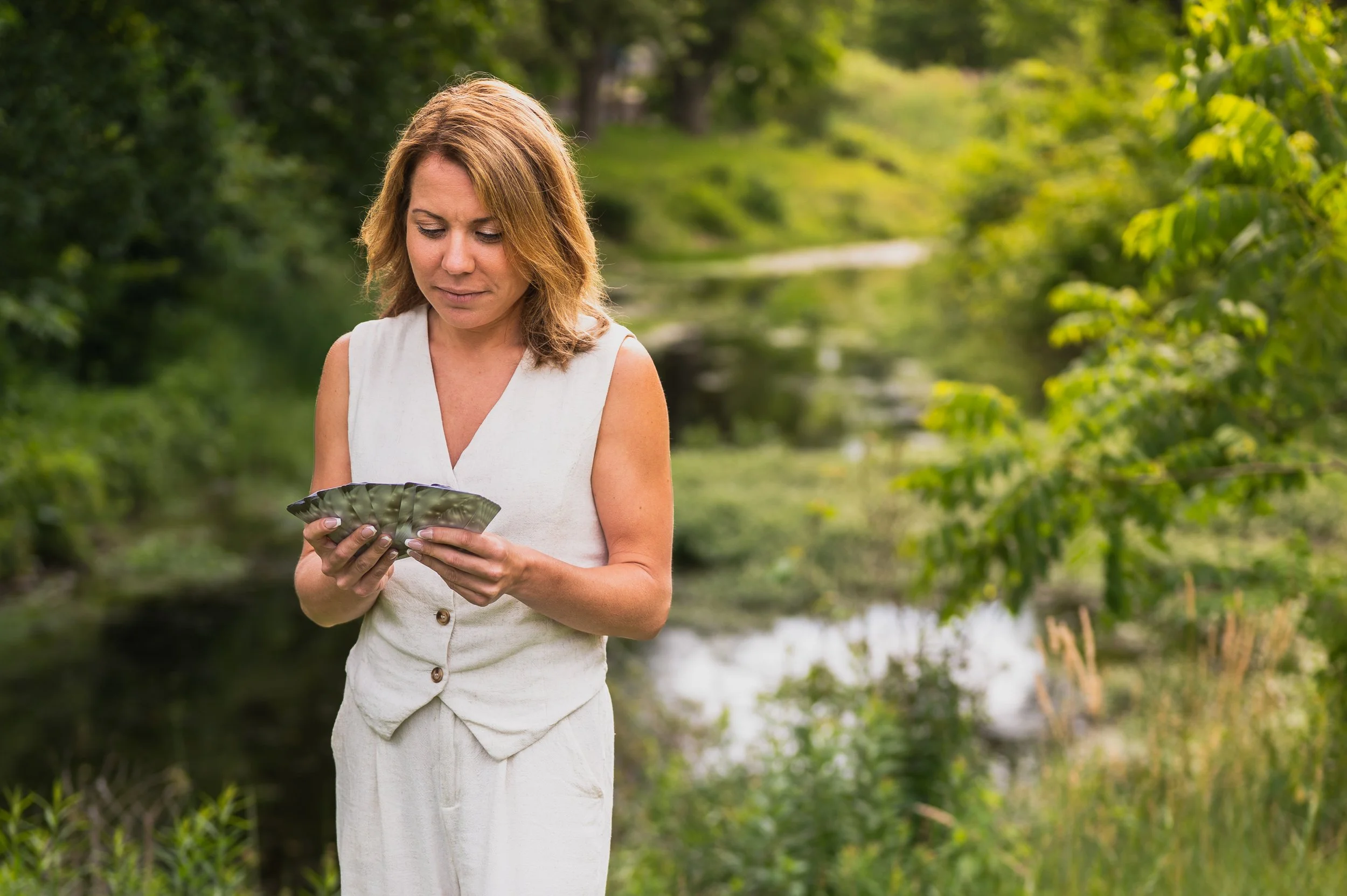 A woman with shoulder-length light brown hair wearing a sleeveless white dress standing outdoors by a river, holding a fan of playing cards, with green trees and water in the background.