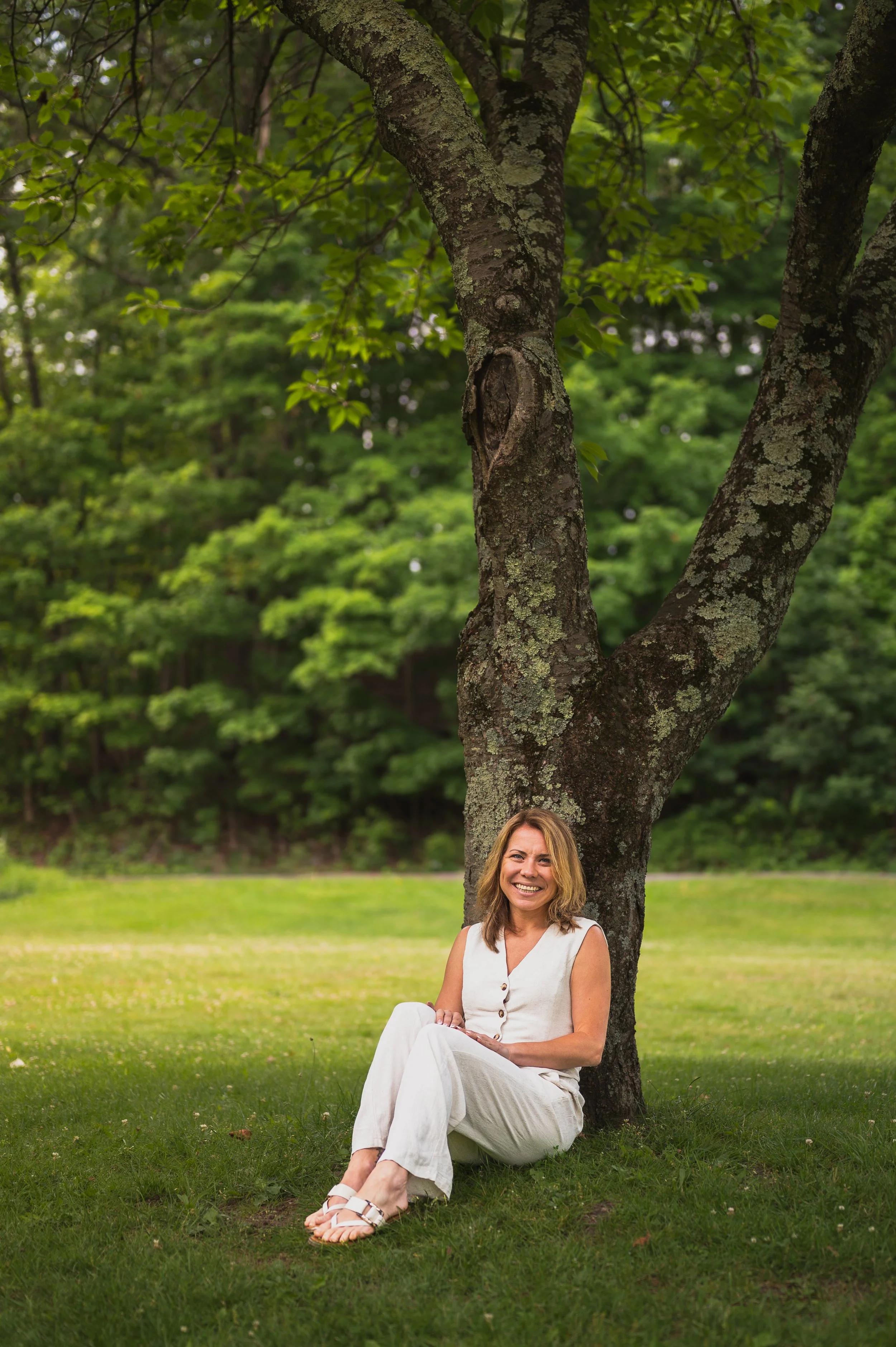 Women sitting under tree the tree has green leaves and grayish bark the women is in a linen suit outfit with short blonde hair the women is smiling