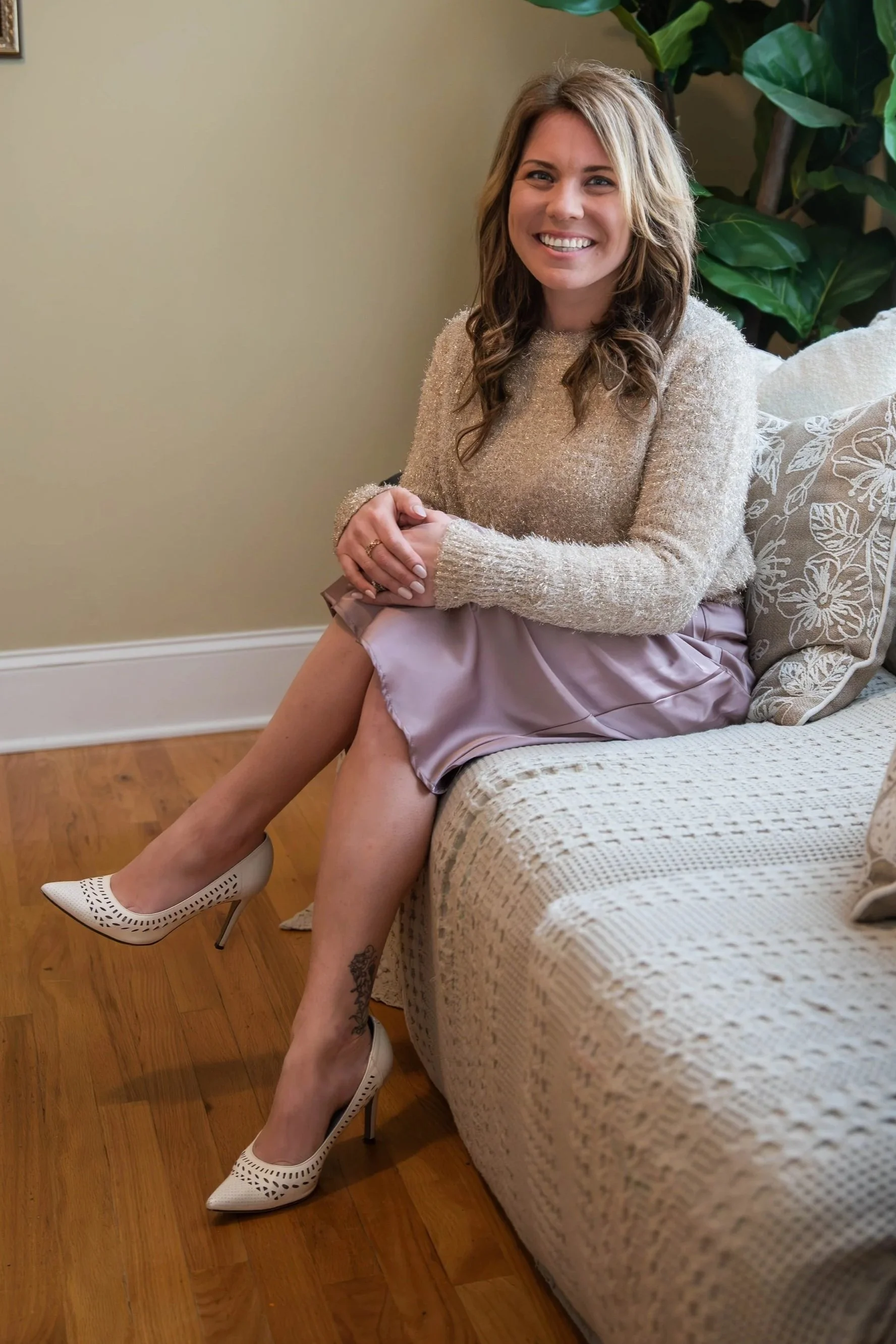 A woman sitting on a sofa, smiling with her hands clasped, wearing a beige sweater, a lavender skirt, and white high heels, with a tattoo on her ankle and a large green plant behind her.