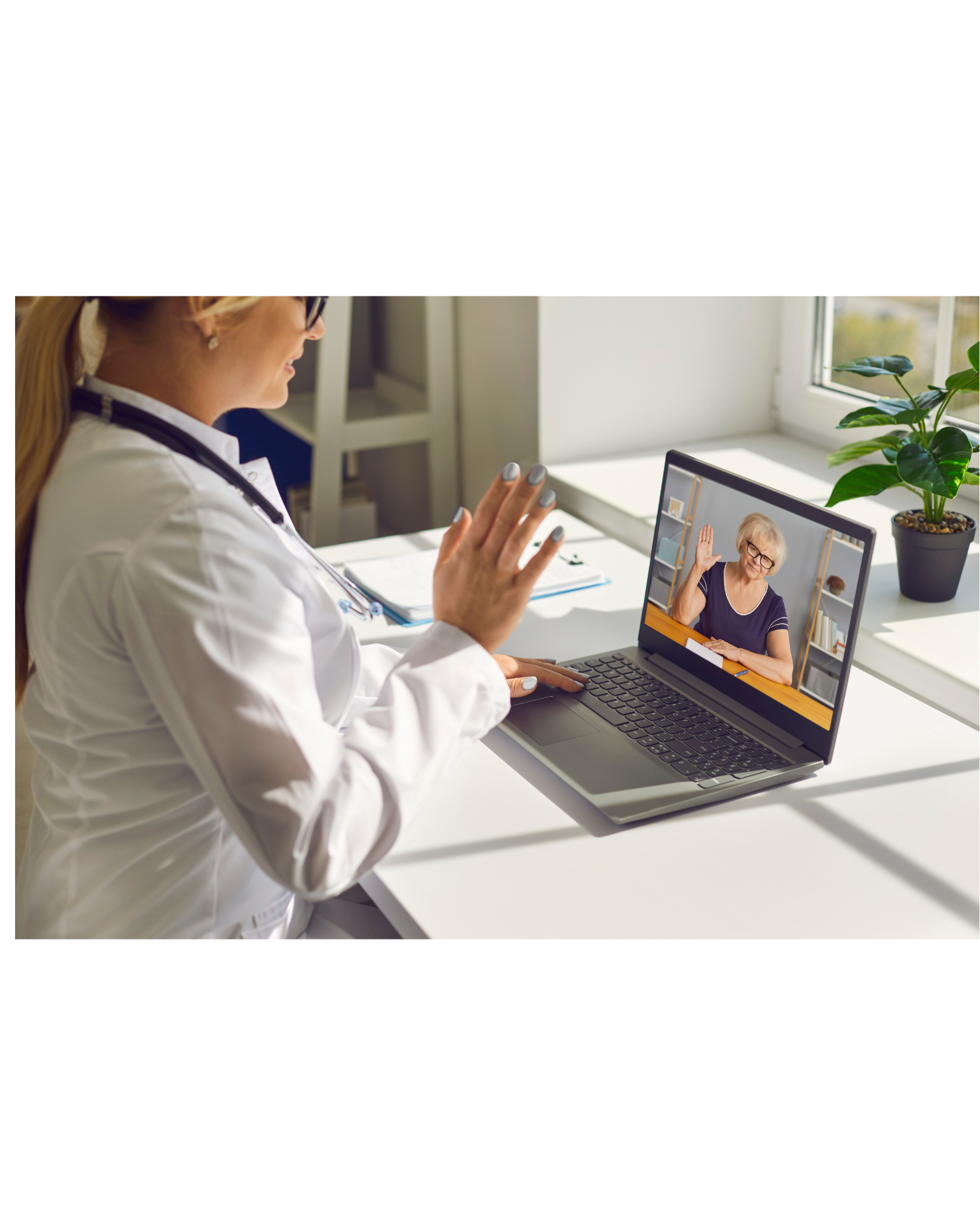 A female doctor in a white coat and stethoscope on her neck is waving at an elderly woman on a video call on her laptop, sitting at a white desk using telehealth.