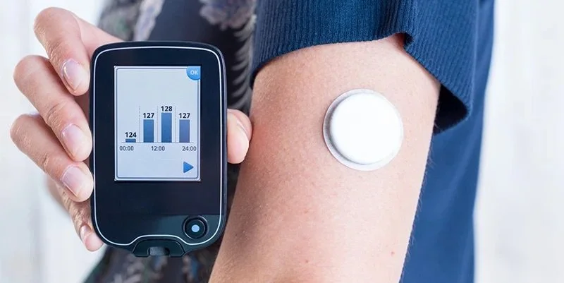 A person holding a blood glucose monitor showing a graph and numbers, with a continuous glucose monitor sensor on their arm.