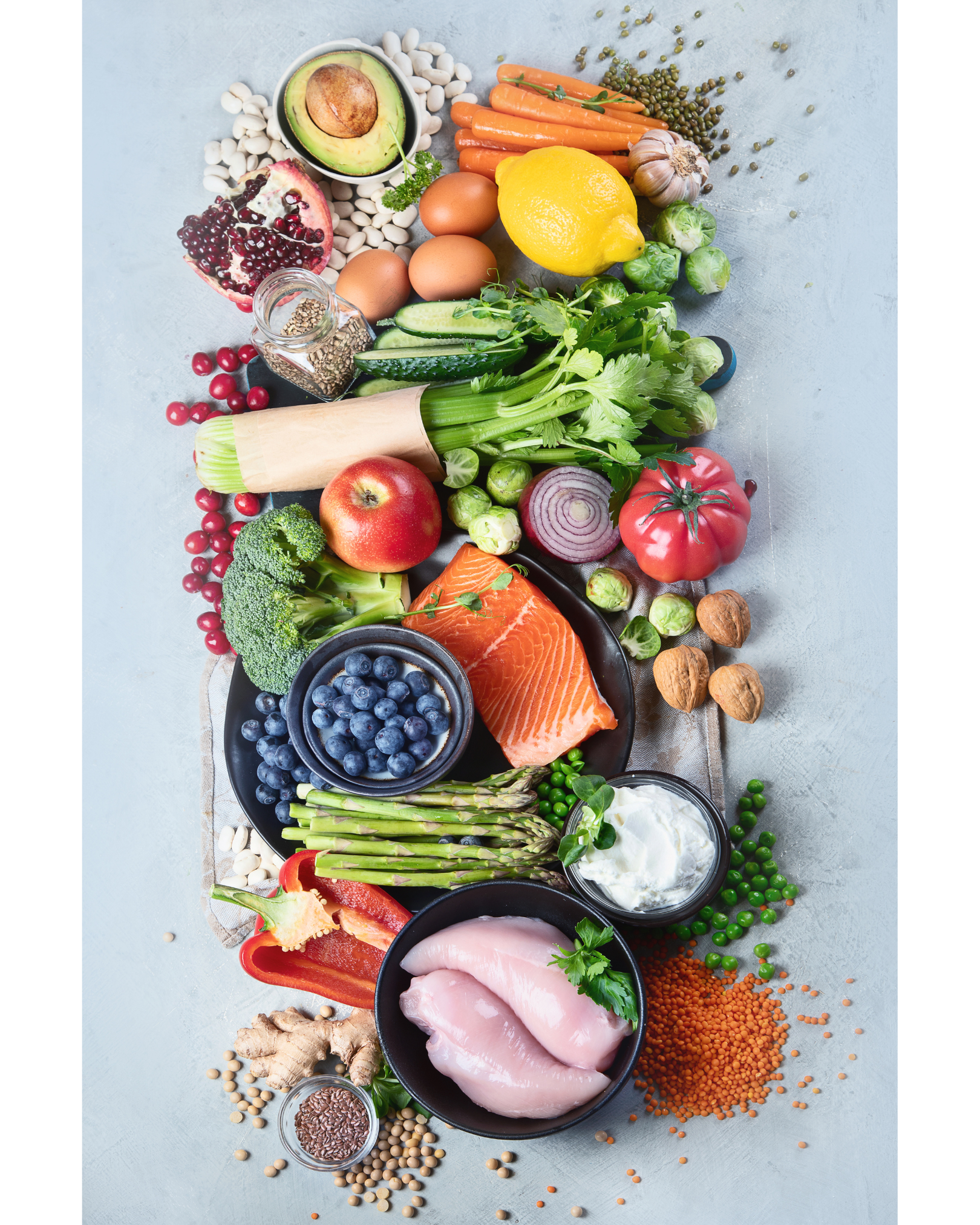 A variety of fresh vegetables, fruits, proteins, and grains arranged on a flat surface, including avocado, carrots, garlic, greens, apple, blueberries, chicken, and lentils.