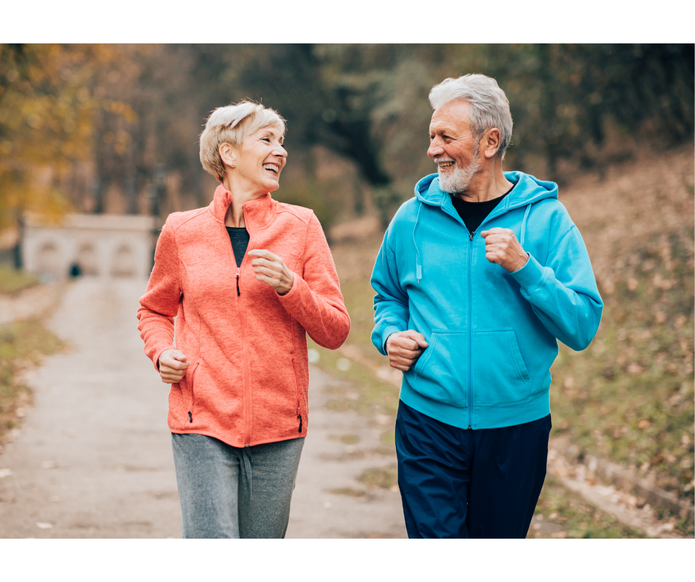 An elderly couple jogging outdoors on a leaf-covered path, smiling and enjoying each other's company.