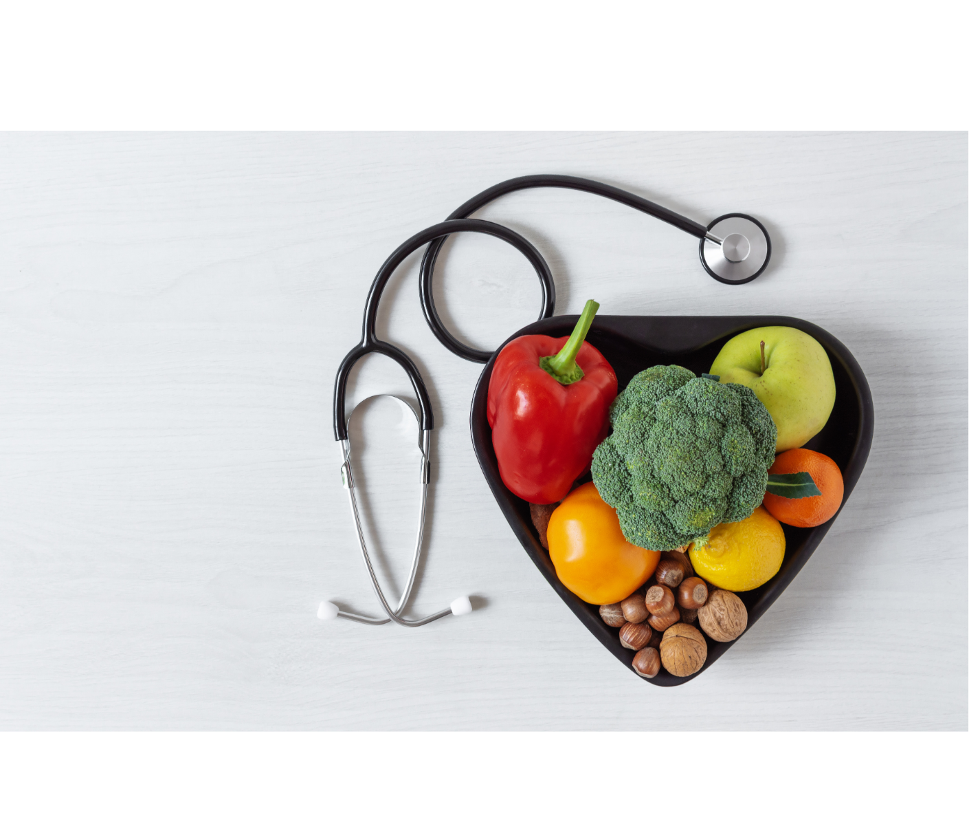 Heart-shaped black bowl filled with colorful fruits and vegetables, including a red bell pepper, green apple, broccoli, yellow bell pepper, orange, lemon, nuts, and hazelnuts, with a stethoscope resting on a white surface nearby.