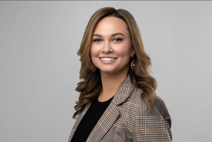 Founder of Balanced Blood Sugar Center , Kristen Leonardes. A young woman with wavy light brown hair and earrings, wearing a plaid blazer and black top, smiling at the camera against a plain gray background.