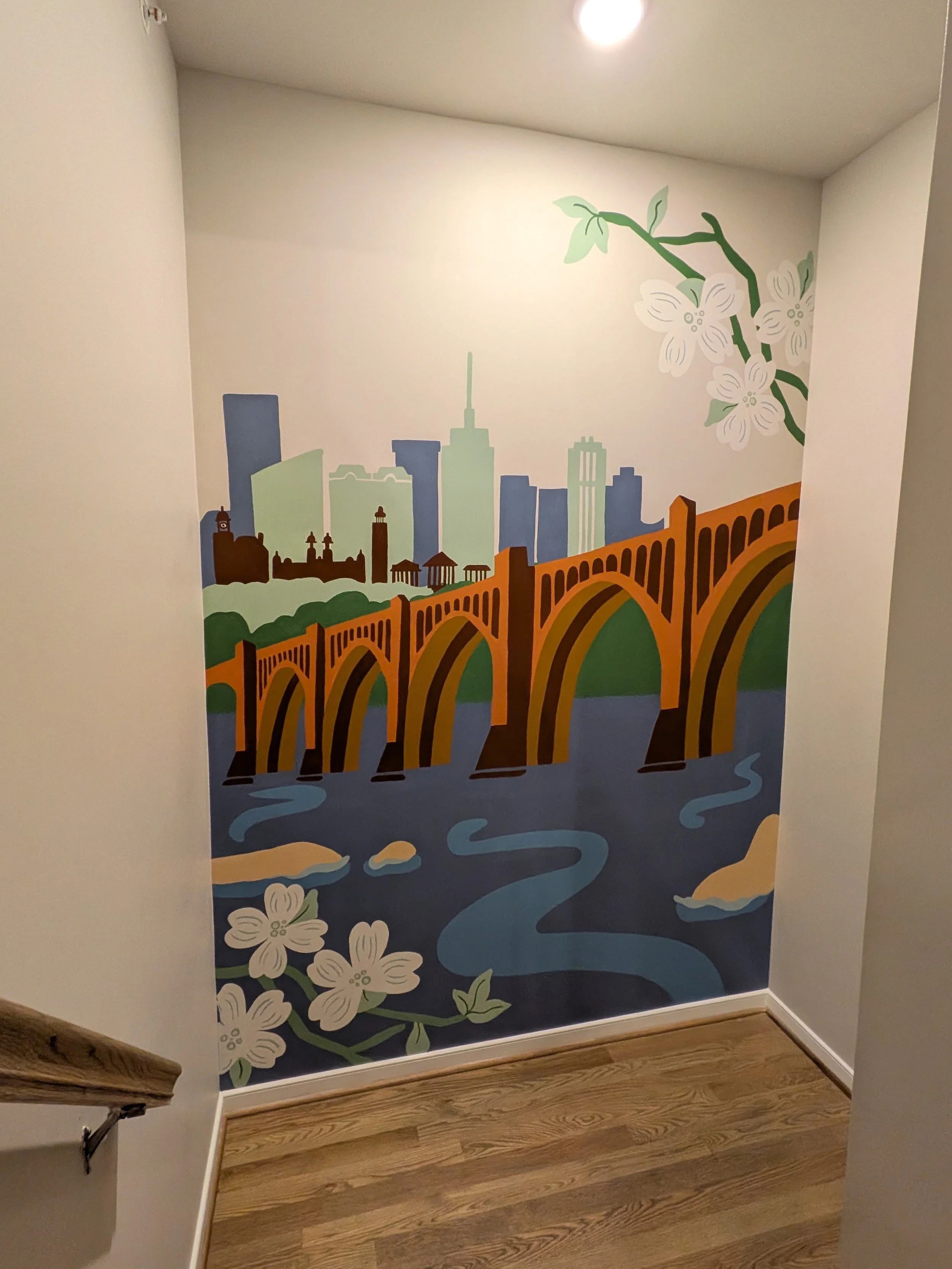 Colorful mural of a city skyline with high-rise buildings, an arched bridge over water, and some flowering branches on the wall.