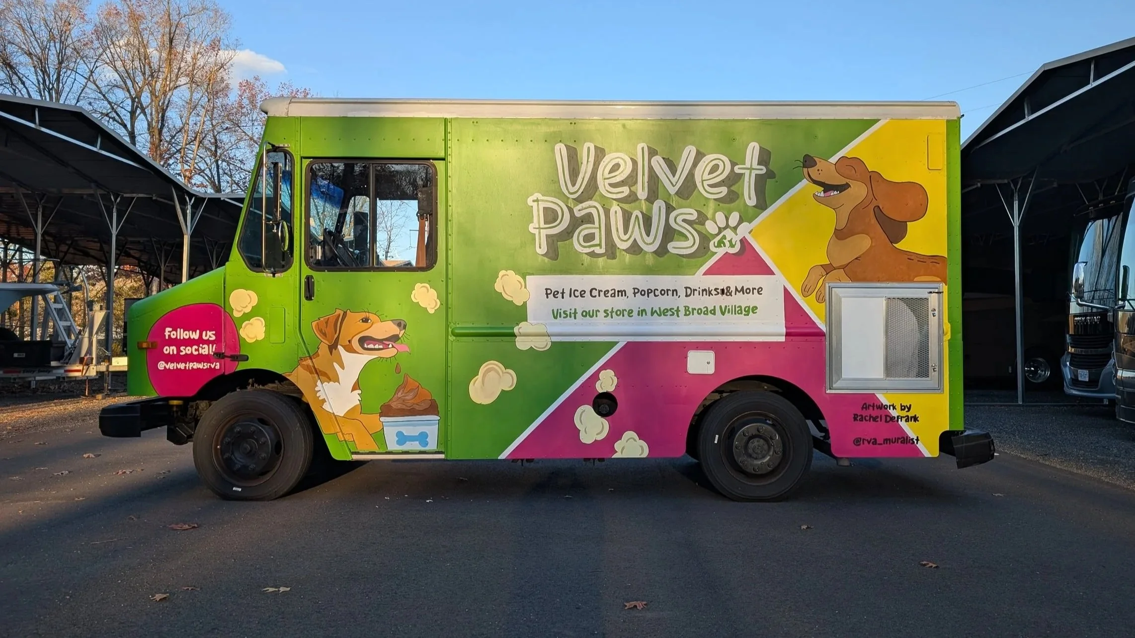 A colorful food truck named Velvet Paws featuring a cartoon dog, with green, pink, and yellow sections. The truck advertises pet ice cream, popcorn, drinks, and more, with social media information and artwork credit to Rachel Derank.