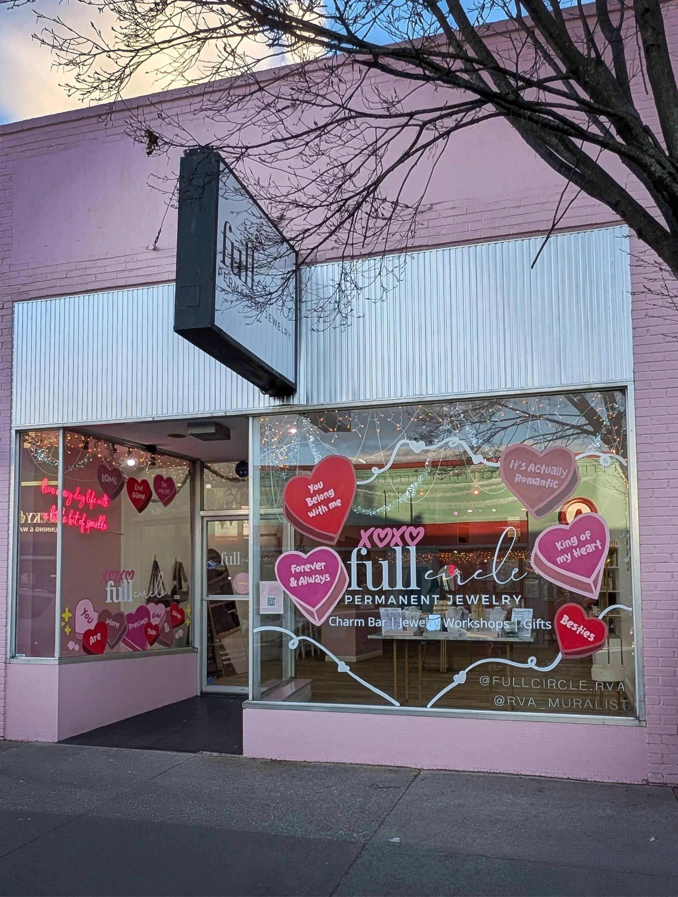 The storefront of Full Circle Permanent Jewelry, decorated with pink and red heart-shaped signs with messages like "You Belong with me" and "Forever & Always." The store has a large window with the shop's name and various signs advertising charm bar, jewelry workshops, and gifts. Christmas lights are strung across the window, and a tree with leafless branches is visible above.