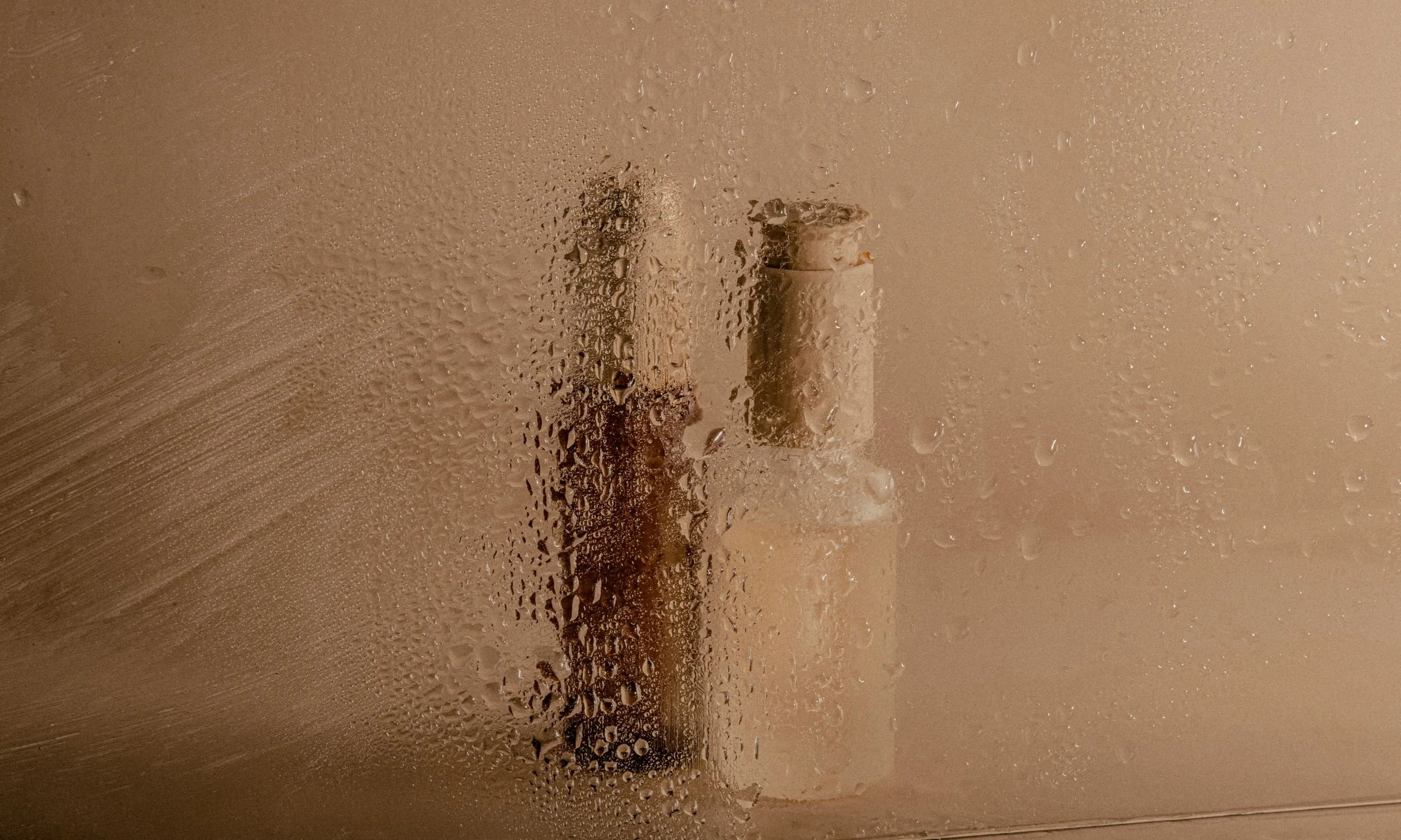 View through a foggy or steamy glass shower door, with condensation and water droplets covering the surface. Behind the glass, there are two bottles, possibly shampoo, conditioner, or body wash, placed on a shower shelf or ledge.