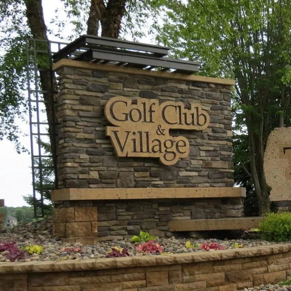 Stone monument sign for a golf club and village, with trees and landscaping in the background.