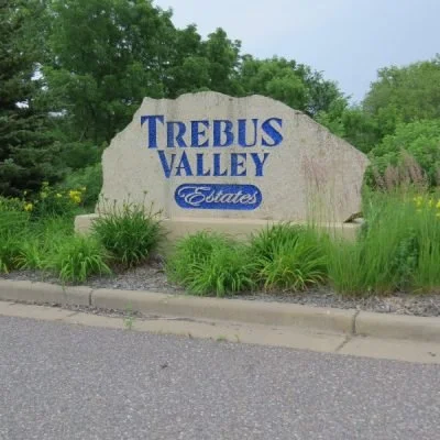 Large stone sign for Trebus Valley Estates with blue lettering, surrounded by green plants and trees.