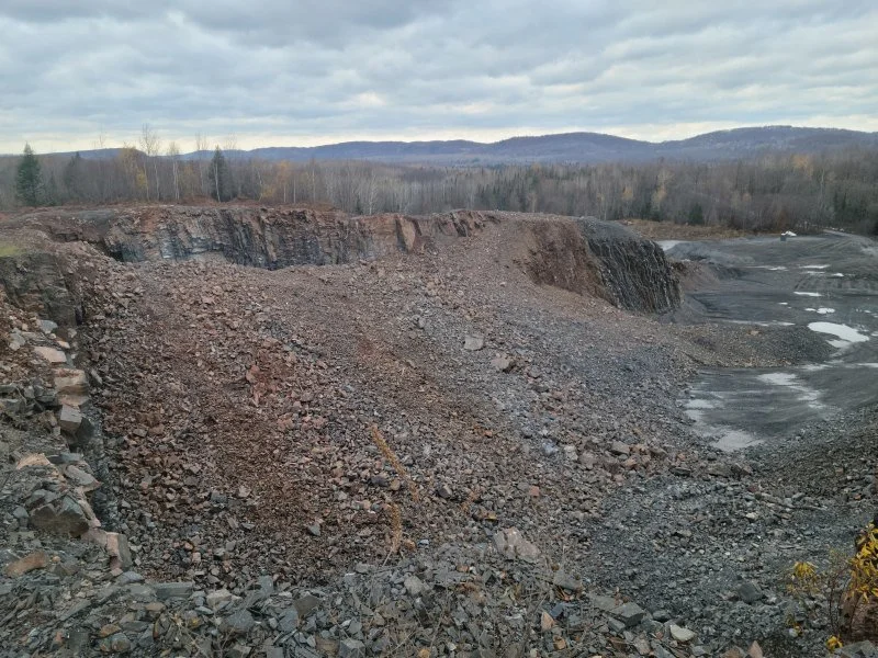 A large open-pit quarry with exposed rock and gravel, surrounded by distant trees and hills under a cloudy sky.