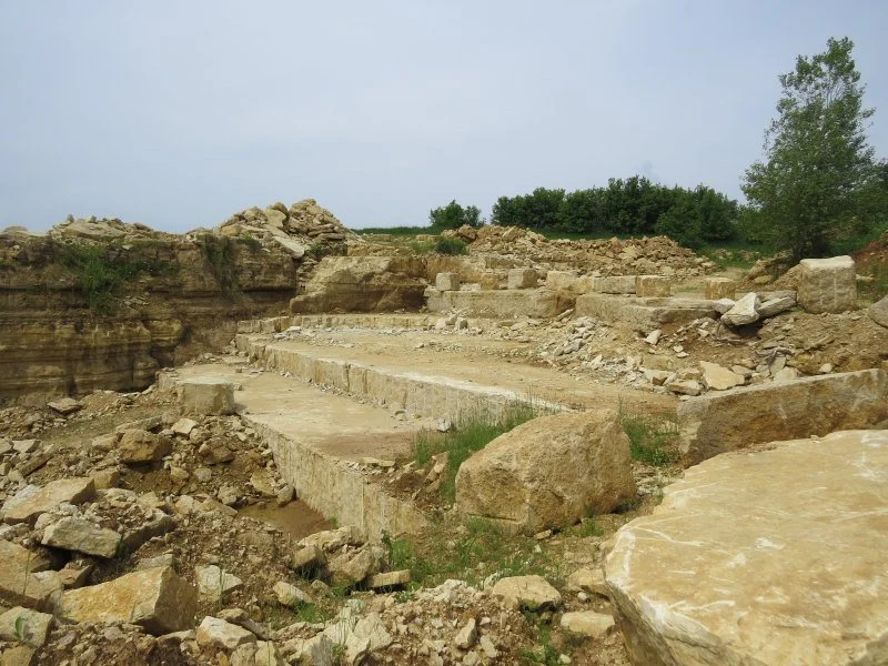 Archaeological site with stone ruins and broken rocks under an overcast sky, surrounded by greenery.