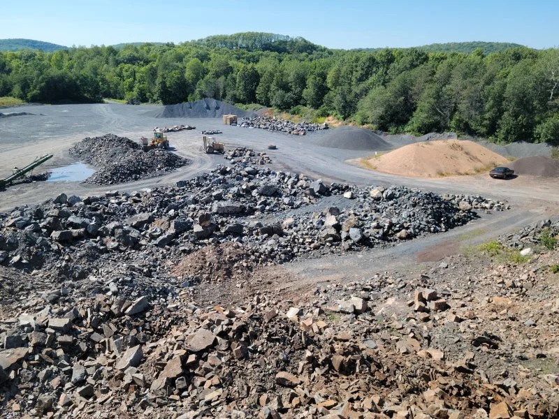 Construction site in a quarry with piles of gravel and rocks, heavy machinery, and a vehicle, surrounded by greenery and trees under a clear blue sky.