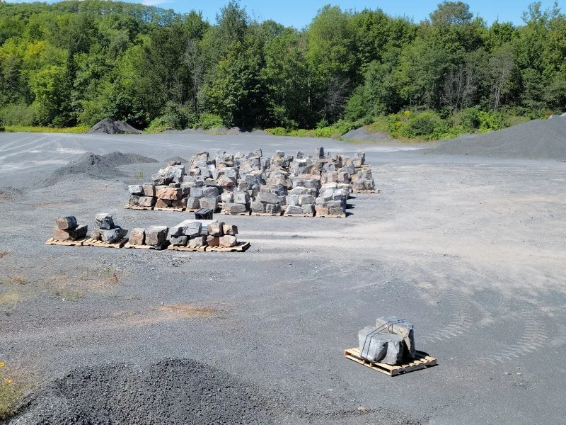 Piles of large rocks and stones on pallets in an open gravel area with trees in the background.