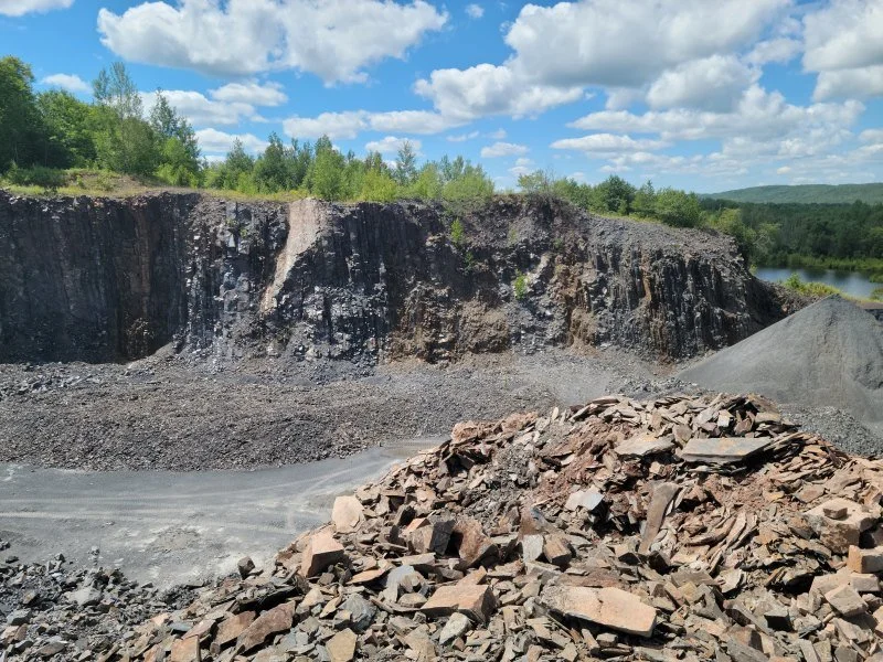 A quarry with a large rocky cliff, piles of gravel, and rocks in the foreground, with trees and a river in the background under a partly cloudy sky.