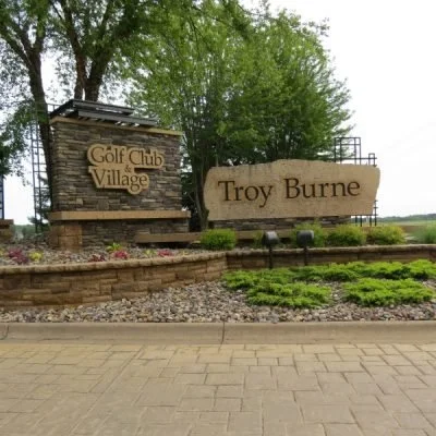 Entrance sign for Golf Club Village and Troy Burne, surrounded by greenery and landscaped with flowers and shrubs.