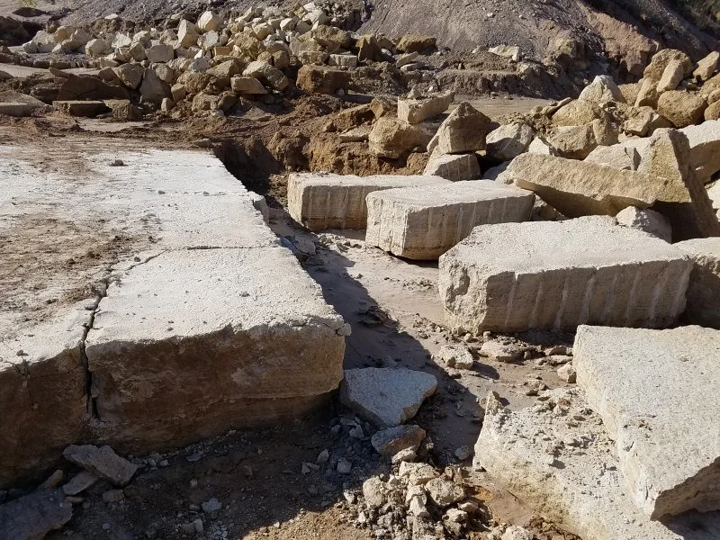 Large concrete blocks and rocks piled along a construction or excavation site, with a partially paved surface and rocky hillside in the background.
