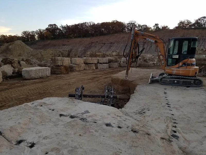 Construction site with an orange mini excavator digging into the ground, surrounded by large stone blocks and dirt, with trees in the background.