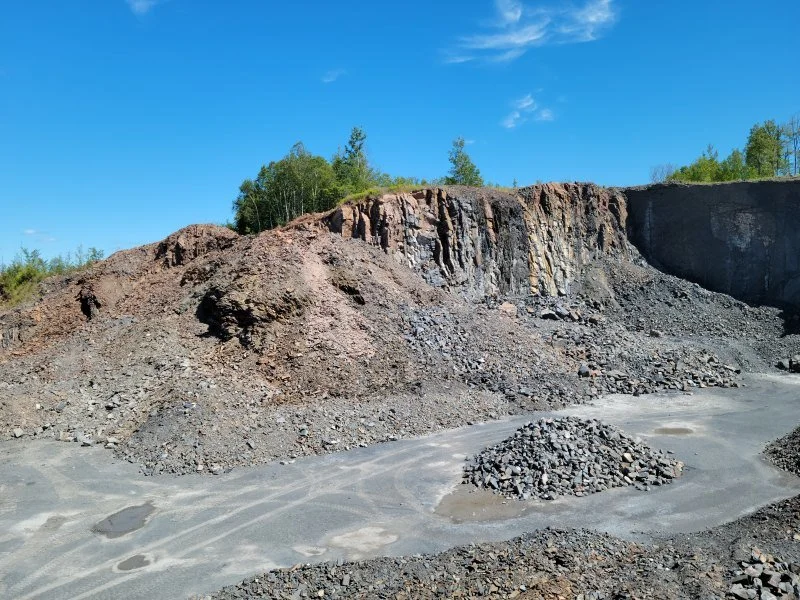 A large open-pit mine with exposed rock faces, piles of mined materials, and a partly gravel surface under a clear blue sky.