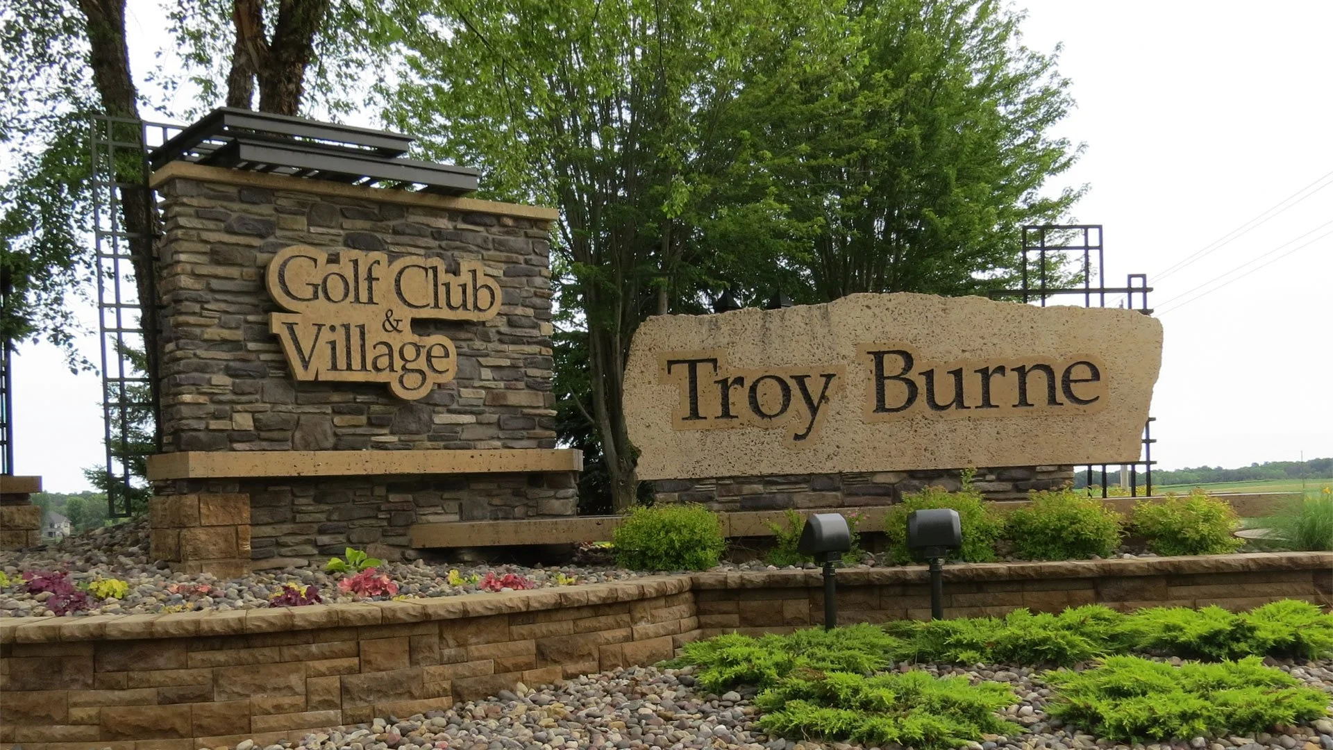 Entrance signage at Troy Burne Golf Club & Village with stone and concrete structures, landscaping, and trees in the background.