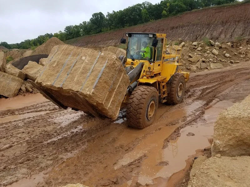 A yellow Volvo front loader moving large rocks in a muddy construction site.