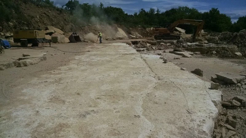 Construction site with broken concrete slabs, an excavator, and a worker wearing a yellow safety vest and helmet