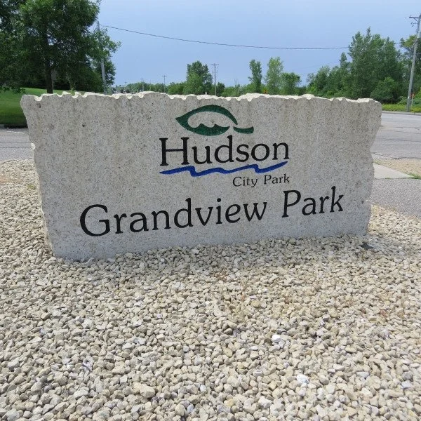 Large stone sign with the text "Hudson City Park Grandview Park" and a fish symbol at the top, situated in a gravel area beside a road.