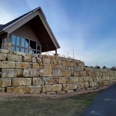 Residential house with large windows and a sloped roof, built on a stone retaining wall, with a driveway and grassy area in the foreground under a blue sky.