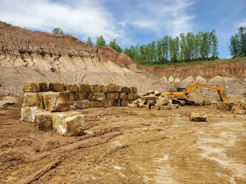 Construction site with an excavator and a partially built stone wall on muddy ground, with a dirt hill and trees in the background.