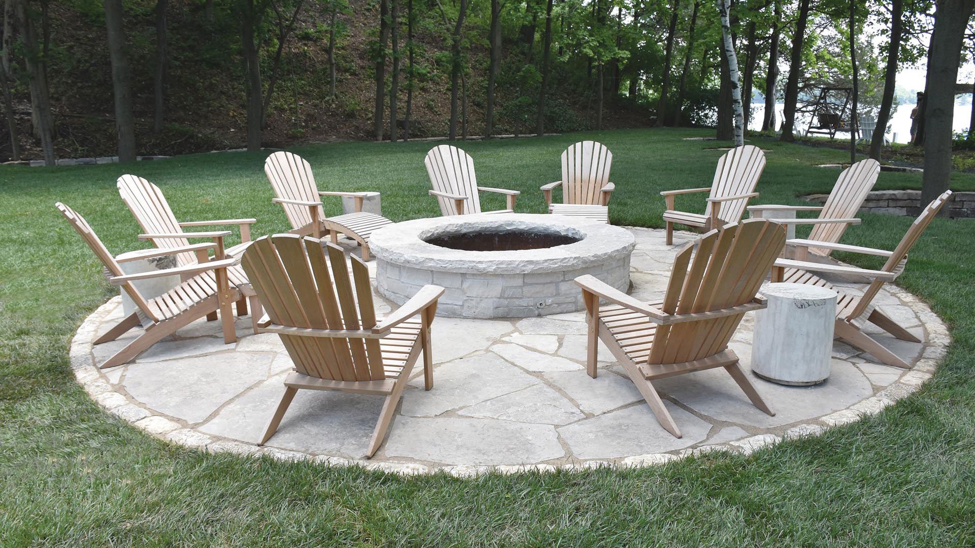 A circular outdoor seating area with wooden chairs arranged around a central stone fire pit on a stone patio, in a grassy backyard with trees and a lake in the background.
