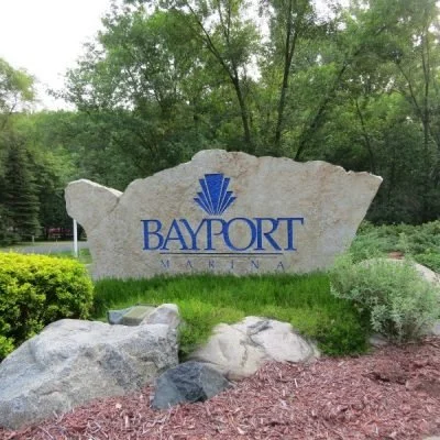 A large stone sign with the word "BAYPORT" and a stylized blue logo, surrounded by greenery and smaller rocks.