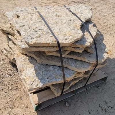Stack of large, rough, irregular stone slabs secured with black straps on a wooden pallet on sandy ground.