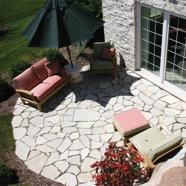 A view of a stone patio outside a house, with outdoor furniture including a bench, two chairs, and an ottoman with red patterned cushions, a large green umbrella, and a sliding glass door.