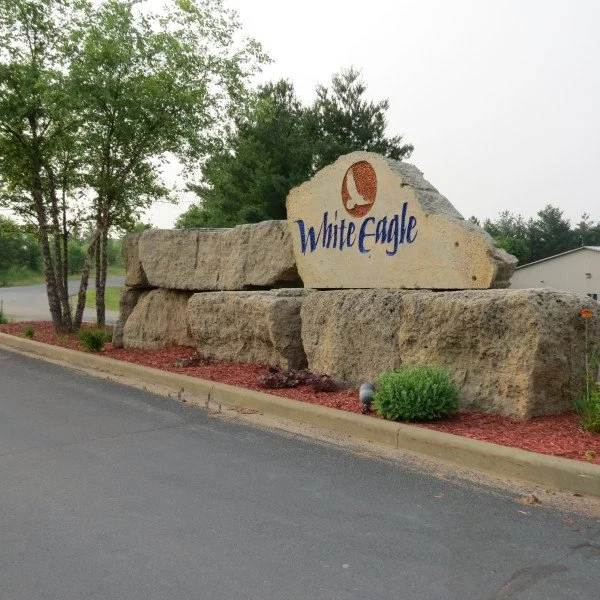 Large rocks with a sign reading 'White Eagle' and a logo of a white eagle head, surrounded by trees and bushes, along a paved road.