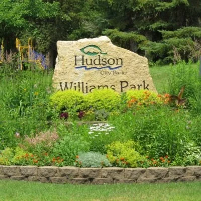 A large stone sign reading 'Hudson City Park Williams Park' with green trees and flowering plants surrounding it.