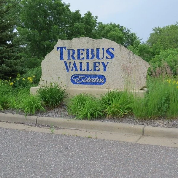 Large stone sign with blue text reading 'Trebus Valley Estates' surrounded by green plants and trees.
