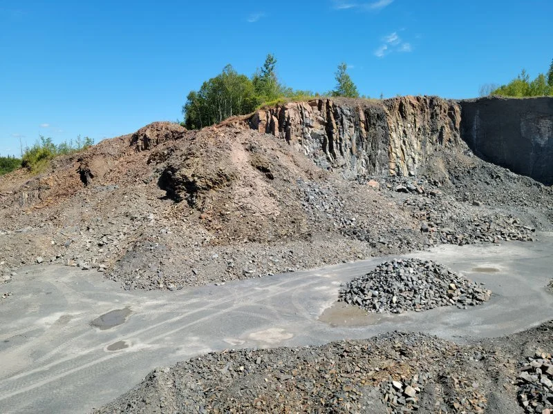 A landscape of an open-pit quarry showing exposed rock layers, a pile of gravel, and a dirt mound under a clear blue sky.
