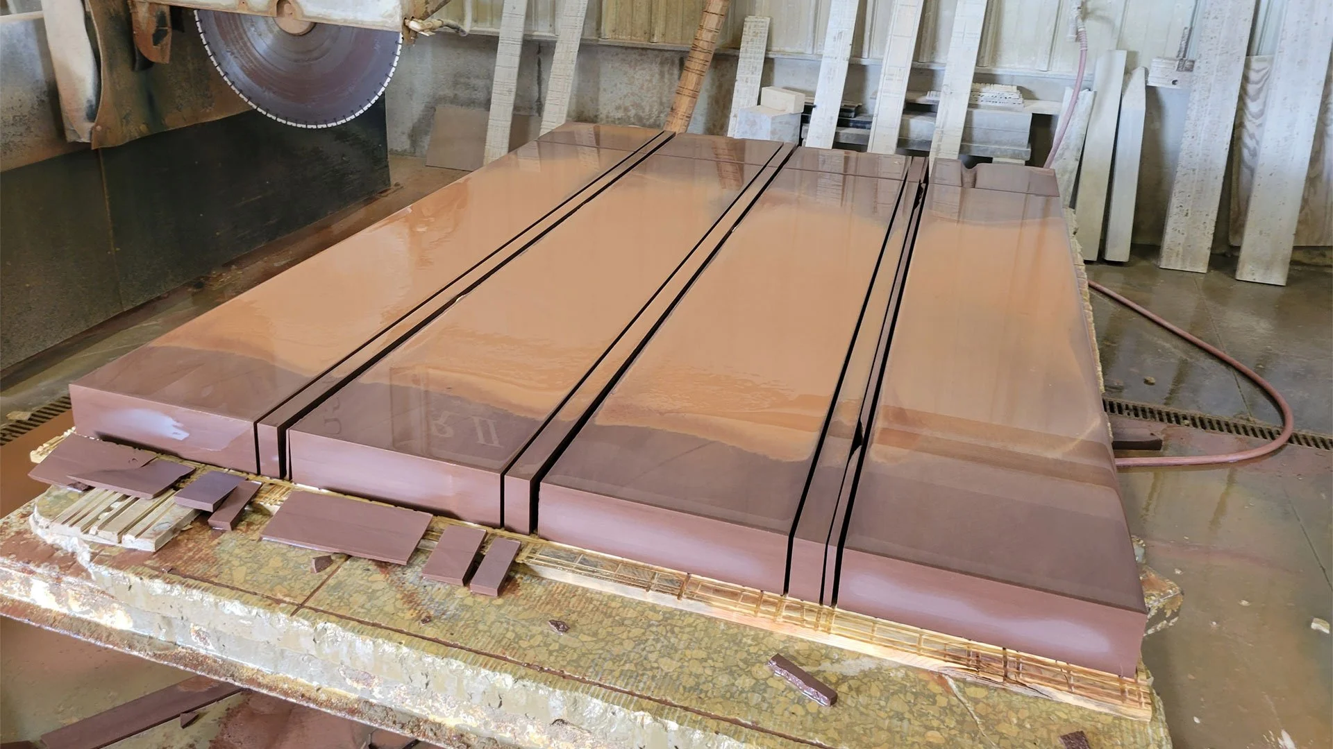 Large wooden workshop table with polished wood panels, black lines marking sections, and smaller wood pieces nearby; background includes woodworking tools and materials.