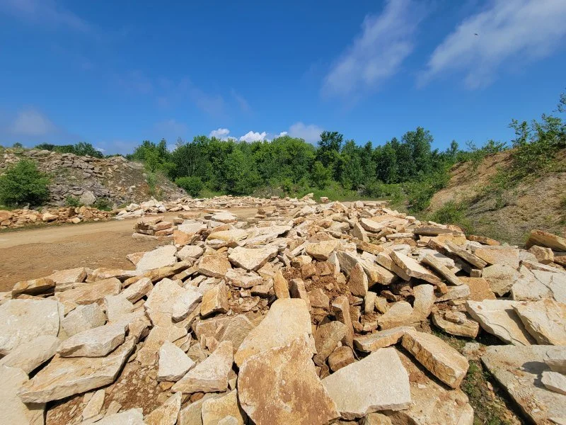A landscape scene showing a rocky area with scattered stones in the foreground, a dirt path, and green trees under a blue sky with some clouds.