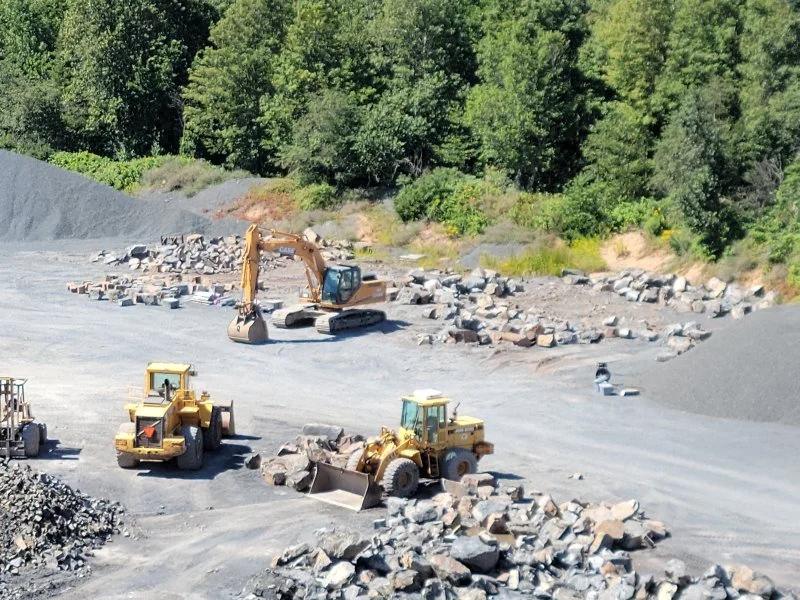 Construction site with bulldozers and an excavator pushing rocks and gravel, surrounded by greenery and trees.