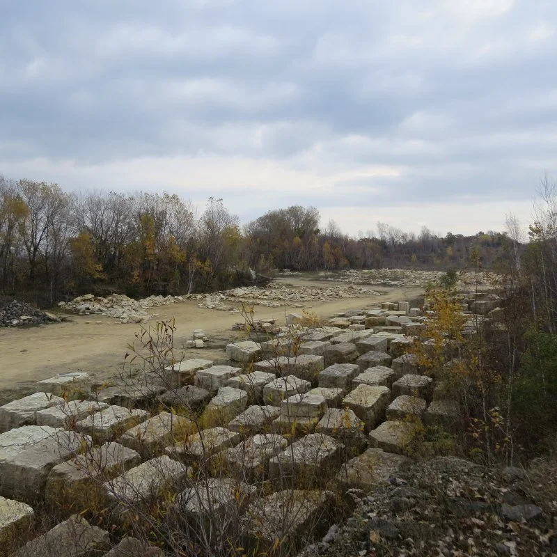 A riverbed with large, rectangular stone blocks along the banks and smaller stones scattered across the dry, sandy area. Trees with sparse autumn foliage line the perimeter under a cloudy sky.