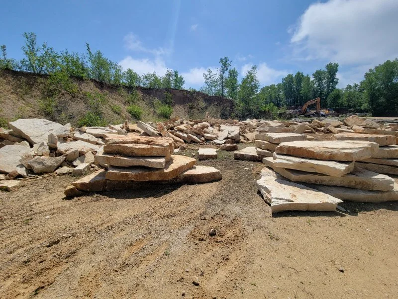 Stacks of flat, light-colored rocks on a dirt surface at a construction or excavation site, with trees and a blue sky in the background.