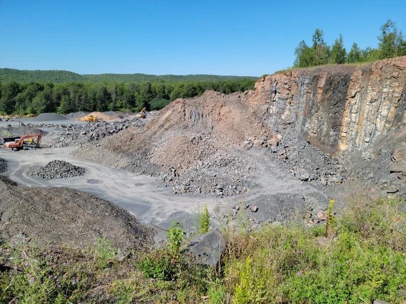 A quarry site with large piles of gravel and dirt, excavation equipment, and a partially exposed rocky cliff face under a clear blue sky.